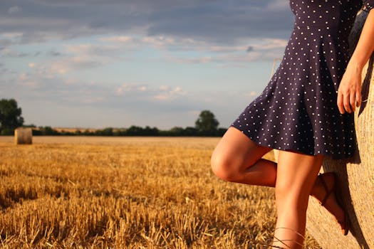 A woman in a polka dot dress leans against a hay bale in a sunlit wheat field.